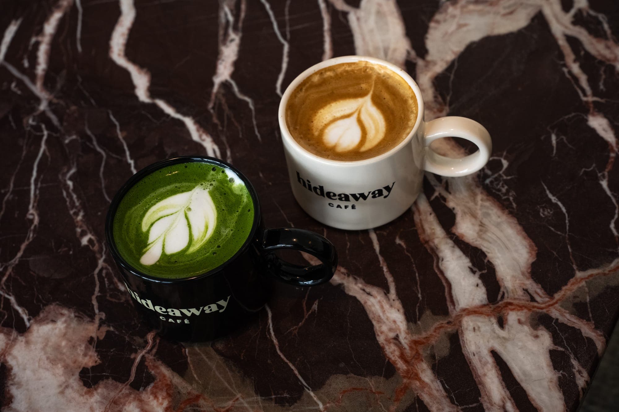 A matcha latte and a cappuccino in hideaway mugs on dark marble