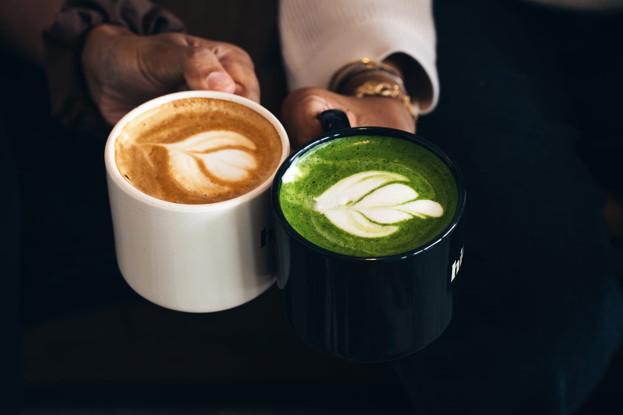 Two hands holding a cappuccino in a white hideaway mug and a matcha latte in a black one, both with latte art
