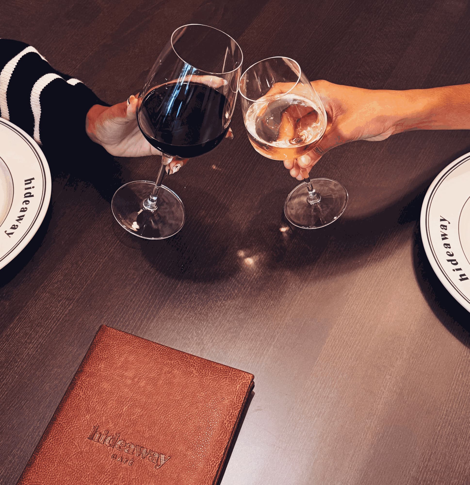 Two hands clinking a glass of red and a glass of white over a dark wooden table, hideaway plates and the leather menu beside them